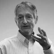 Black and white photo of Geoffrey Hinton. He is speaking with his left hand held up