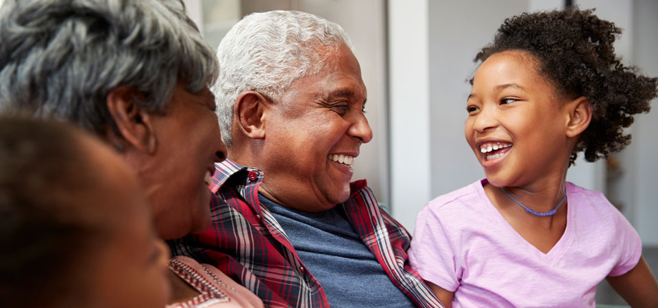 And man and woman sit with their granddaughter. They are smiling. The man is wearing a blue shirt and plaid button down and the granddaughter is wearing a pink tshirt