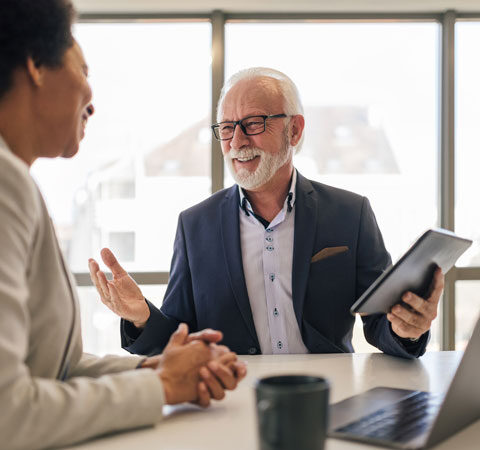 A man with white hair, white beard and glasses talks with a woman. He is wearing a blue suit and is holding a tablet. The woman is wearing a beige suit, has curly hair styled in an afro; her hands are clasped. They are both smiling
