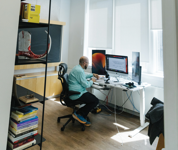 Photo of Alán Aspuru-Guzik sitting in front of a computer at a desk with a bookshelf behind him Photo d'Alán Aspuru-Guzik assis devant un ordinateur à un bureau avec une étagère derrière