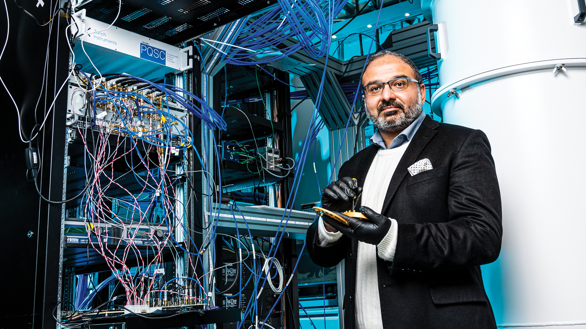 Photo of Irfan Siddiqi standing in front a large computer, wearing black disposable gloves, holding a quantum chip Photo d'Irfan Siddiqi debout devant un grand ordinateur, portant des gants noirs jetables et tenant une puce quantique