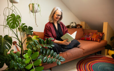 A photo of Kristin Andrews sitting on a rust-coloured couch reading a book. There are a couple of stuffed animals sitting on the top right corner of the couch in the background. A plant is in the bottom-left corner of the photo.	Photo de Kristin Andrews assise sur un canapé couleur rouille, en train de lire un livre. Deux animaux en peluche sont assis dans le coin supérieur droit du canapé, à l’arrière-plan. Il y a une plante dans le coin inférieur gauche de la photo.