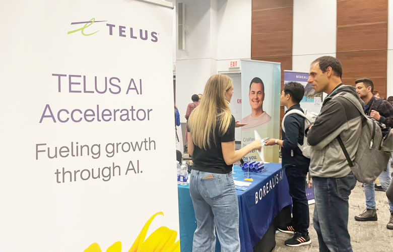 Photo of a TELUS sign in front of a table. There are people standing near the table having a conversation.