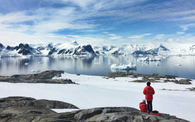 Two people in red jackets on a snowy, rocky terrain overlook a serene Arctic landscape with icy waters and snow-capped mountains under a blue, cloudy sky.