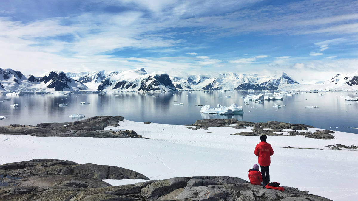Two people in red jackets on a snowy, rocky terrain overlook a serene Arctic landscape with icy waters and snow-capped mountains under a blue, cloudy sky.